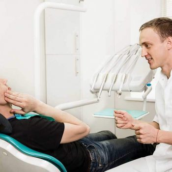 Dentist talking to patient who is in a dental chair and holding his jaw as though he’s in pain.